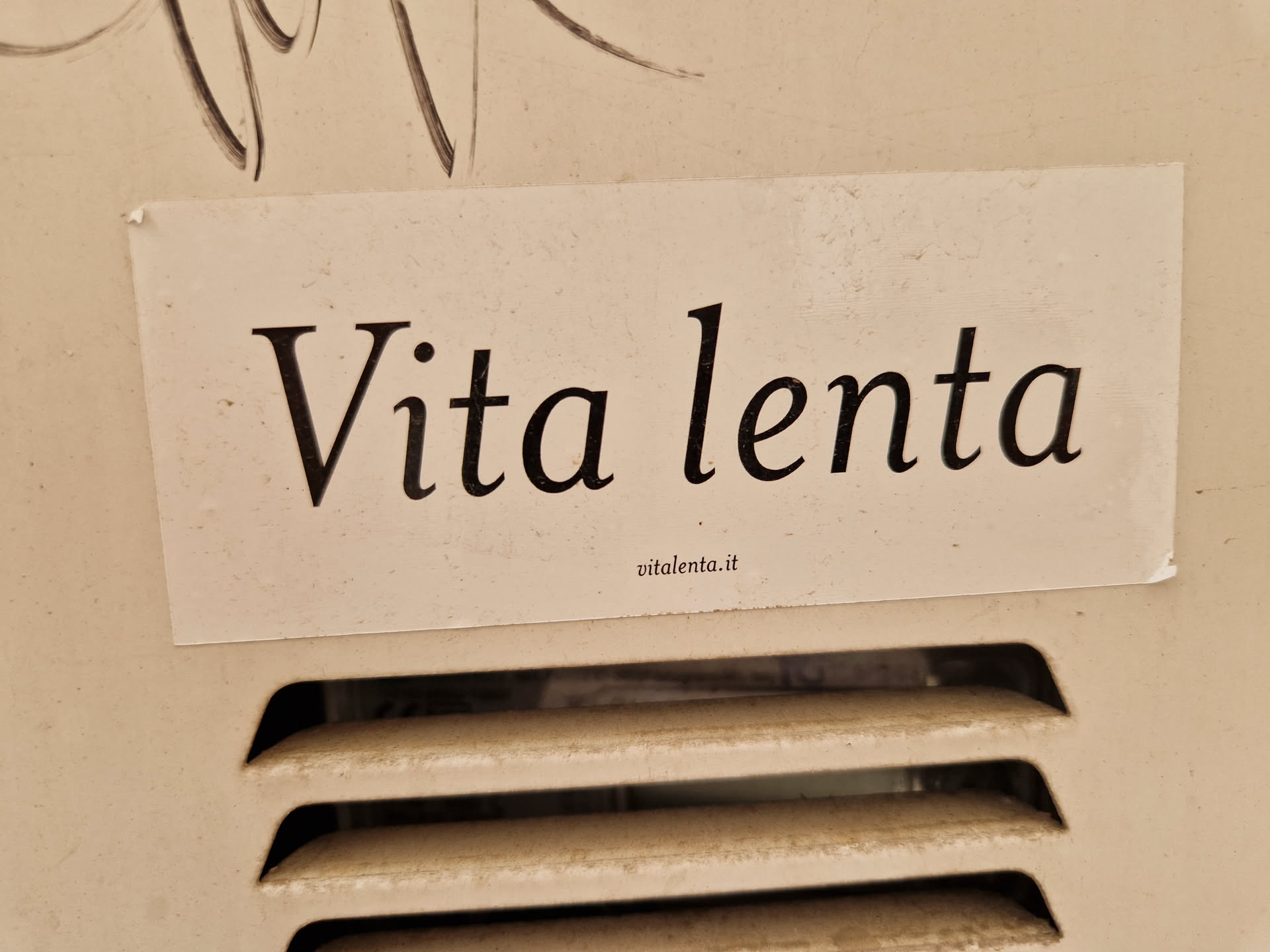 A beige sticker with the text "Vita lenta" (slow life in Italian) on it. The sticker is attached to a beige wall.  There is a small website address visible at the bottom of the sticker.