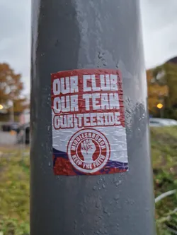 A red and white sticker with a clenched fist in the center, promoting Middlesbrough Football Club. The sticker is affixed to a gray pole outdoors.