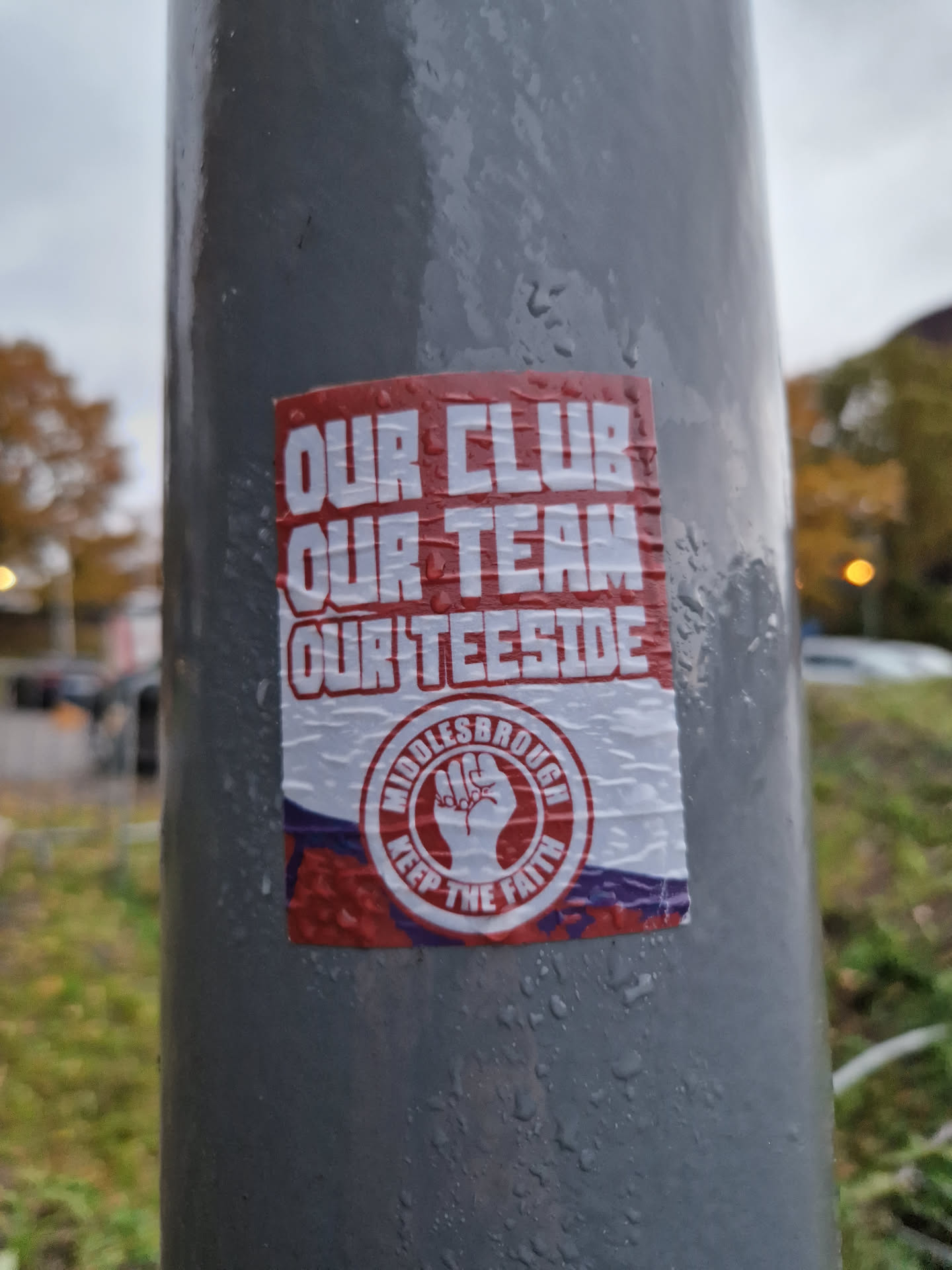 A red and white sticker with a clenched fist in the center, promoting Middlesbrough Football Club. The sticker is affixed to a gray pole outdoors.