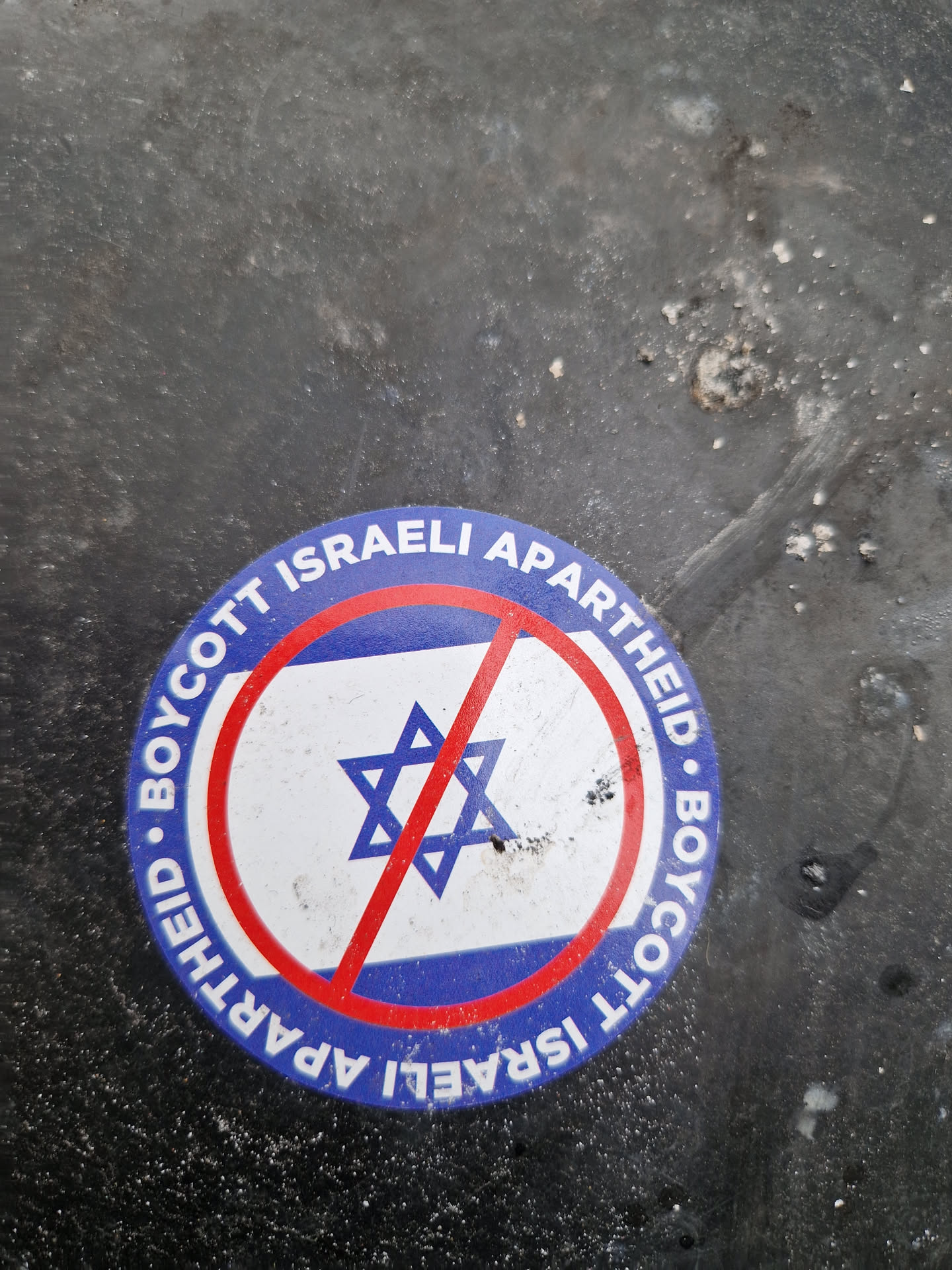 A sticker with a red circle and a line crossing the Israeli flag. The text around the circle says "Boycott Israeli Apartheid" in English.