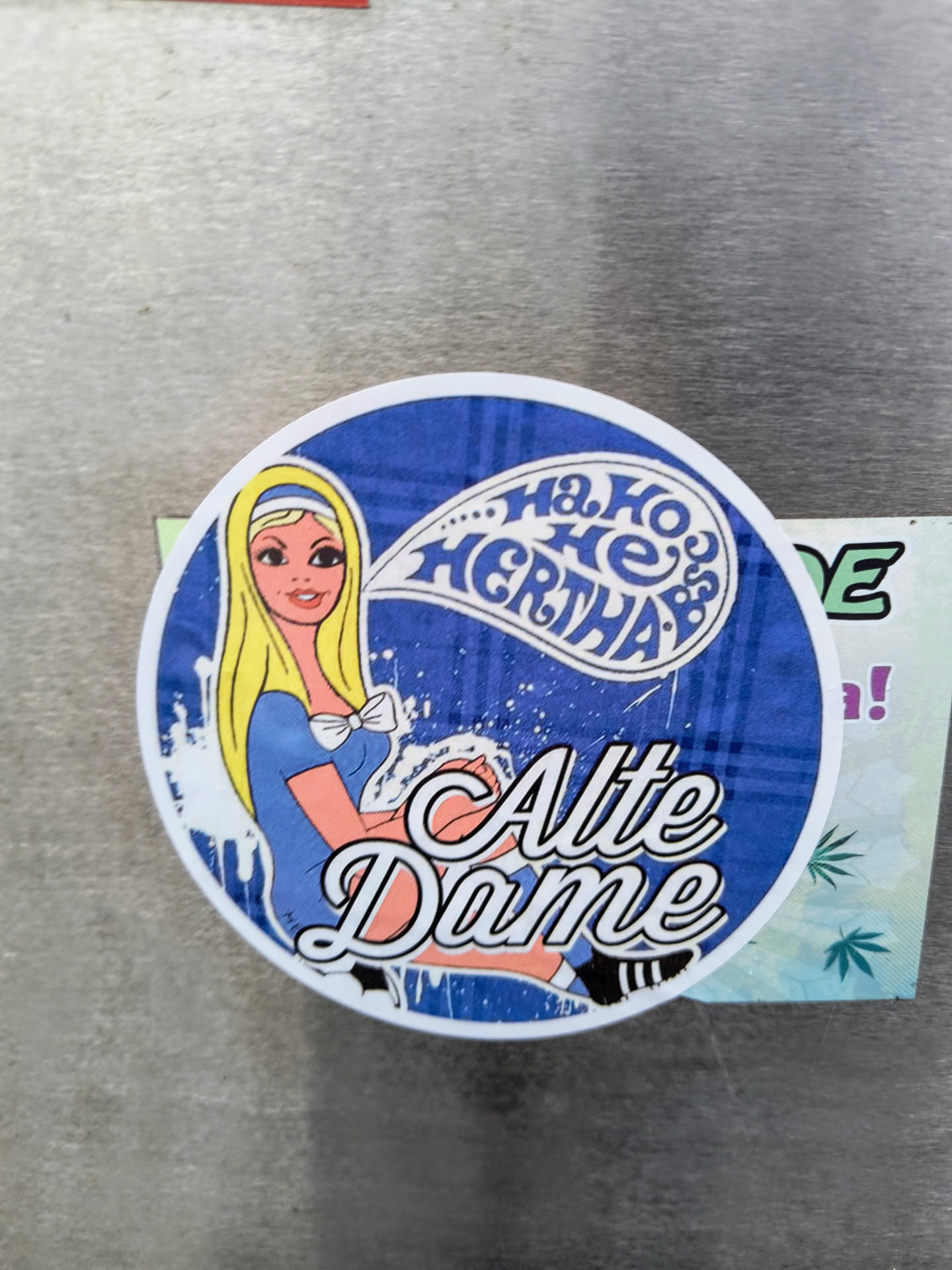 Round sticker with a blue background and a retro-style illustration of a blonde woman in a blue dress. The speech bubble above her says "Ha, Ho, He Hertha." The text "Alte Dame" is prominently displayed below the woman. The sticker has a slightly distressed or vintage look.
