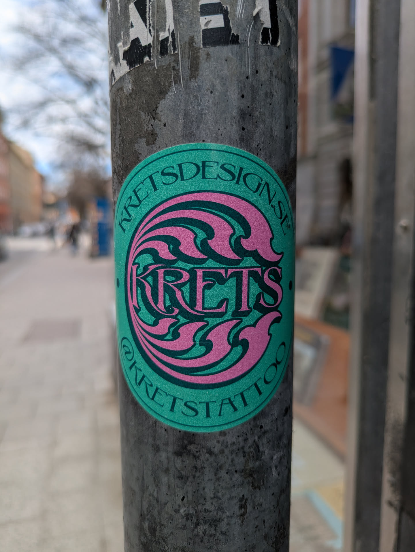 This round sticker features a turquoise background with stylized pink waves and the artist's name 'KRETS' in the center. The artist's website 'KRETSDESIGN.SE' is printed along the top edge, and their Instagram handle '@KRETSTATTOO' is at the bottom. The sticker is affixed to a weathered pole.