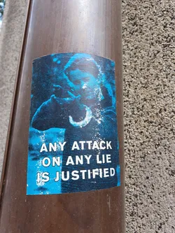 A blue sticker with a picture and a phrase on it. The phrase says: Any attack on any lie is justified.