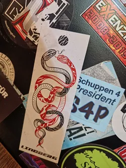 A sticker featuring a minimalist design of two snakes intertwined, one red and one black and white. The background is off-white, and there is a small, dark circle near the top. The word "LINDSBERG" is printed at the bottom.