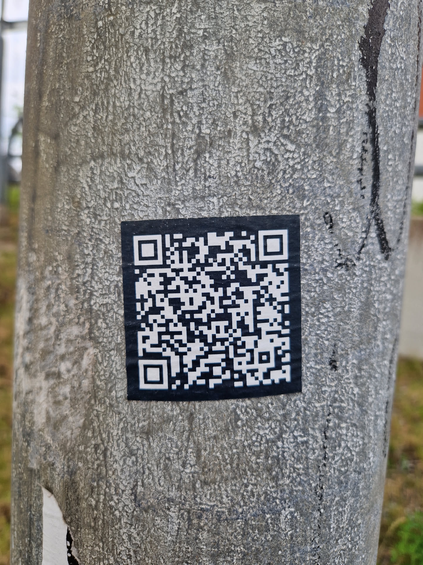 A black and white QR code sticker is affixed to a weathered gray pole.  The sticker is square and the QR code is clearly visible.