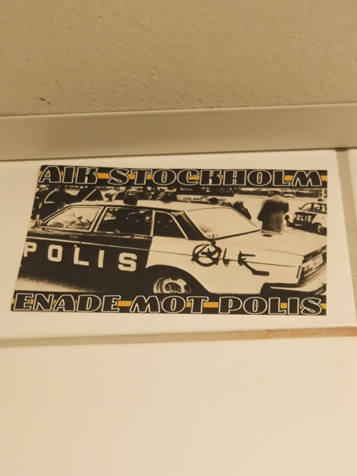 A sticker depicting a black and white image of a police car with graffiti on it. The text on the sticker says "AIR STOCKHOLM" at the top and "ENADE MOT POLIS" at the bottom.  The word POLIS is visible on the car itself. The image appears to be from a demonstration or protest.