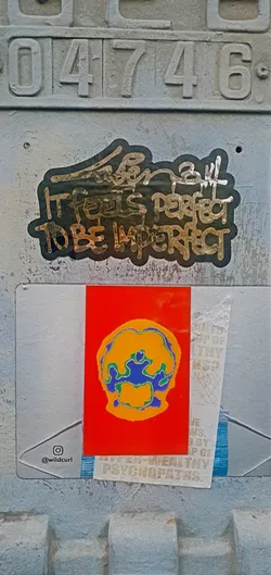 The image features a cluster of stickers adhered to a textured, possibly metallic, surface. At the top, the number "04746" is engraved or stamped. The stickers are layered, with one featuring the phrase "It feels perfect to be imperfect". Another sticker has a vibrant red background with an abstract design in yellow, blue, and green. A ripped sticker reveals partial text like "UP OF HEALTHY," "JE NS, BY IP OF," and "UPPER-WEALTHY PSYCHOPATHS."