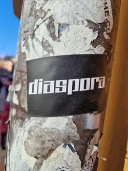 A black sticker with the word "diaspora" in a sans-serif font is affixed to a weathered pole or column.  The surface is covered in remnants of other stickers and shows significant wear.