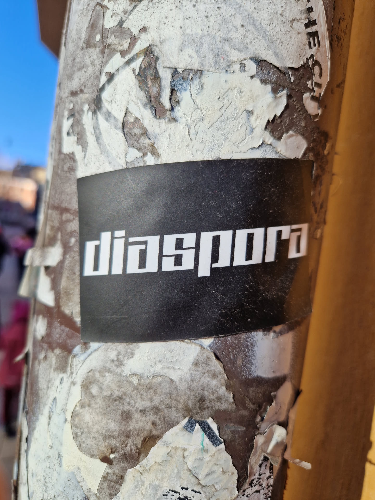 A black sticker with the word "diaspora" in a sans-serif font is affixed to a weathered pole or column.  The surface is covered in remnants of other stickers and shows significant wear.