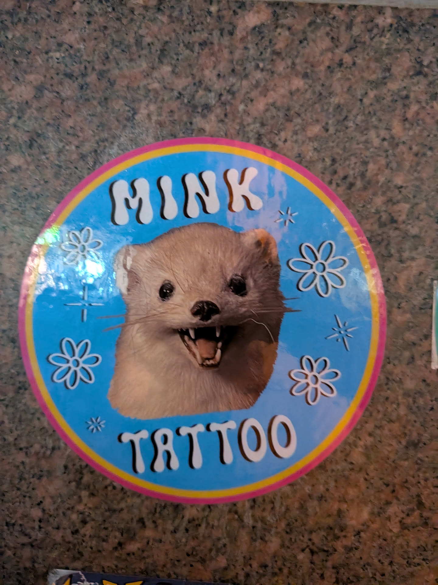 The sticker is circular with a bright blue background. It features a realistic image of a mink's head in the center, with its mouth open, revealing sharp teeth. The mink has dark eyes and light brown fur. Around the mink's head are several white line drawings of flowers and starbursts. The text 'MINK' is prominently displayed in white, bubbly letters at the top of the circle, and 'TATTOO' is similarly displayed at the bottom. The entire design is bordered by a pink and yellow ring.