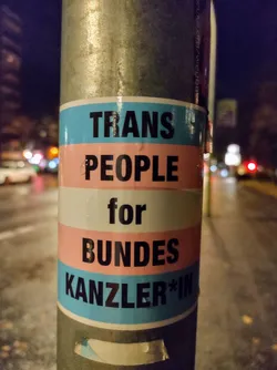 A sticker advocating for transgender people in German politics. The sticker uses the transgender flag colors and the text is in German.