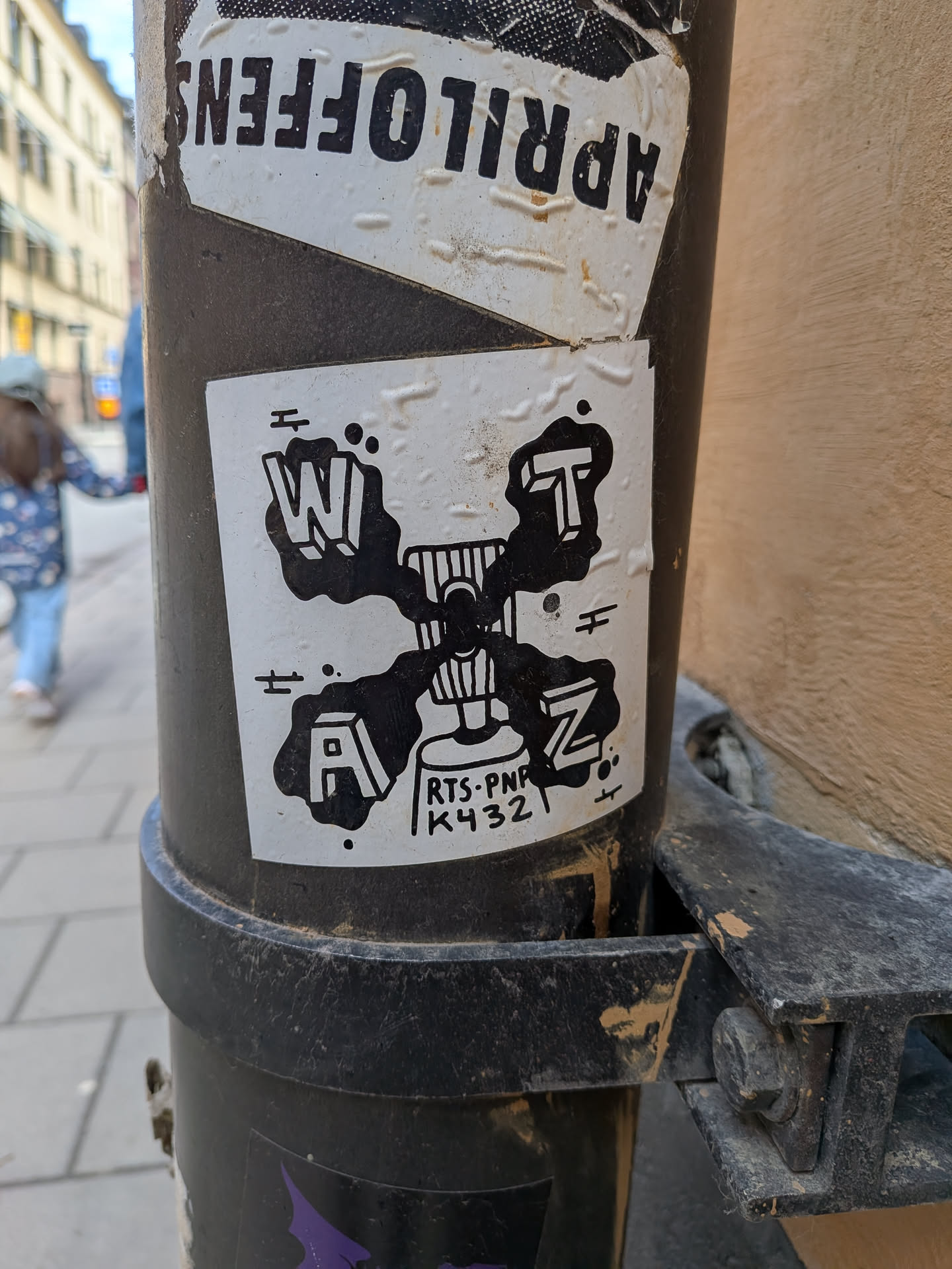 The sticker is square-shaped with a white background and features a bold black design. At its center is an abstract, stylized figure that resembles a person sitting in a chair or a unique lamp, rendered with vertical stripes and a circular head. Surrounding this central figure are four large, block-style letters, each encased in an irregular black 'splatter' or 'drip' shape. The letters are 'W' (top-left), 'T' (top-right), 'Z' (bottom-right), and 'A' (bottom-left). Small horizontal lines with dots are scattered around the design, adding to its graphic quality. At the bottom center, there is smaller, handwritten-style text. The overall aesthetic is monochromatic and graphic, with a distinct street art feel.