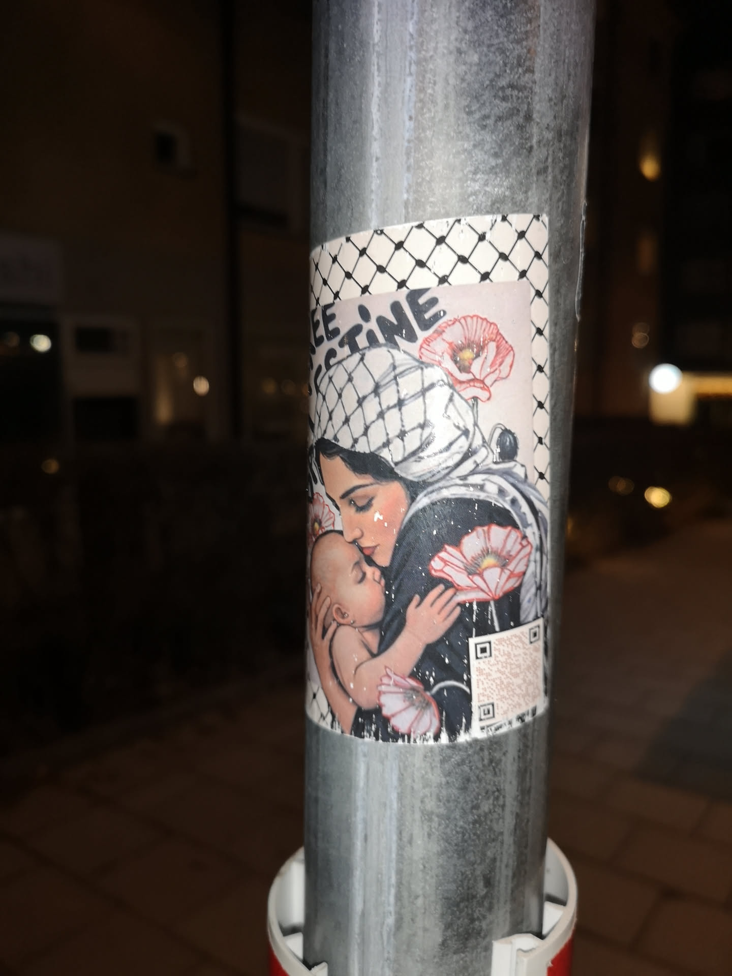 The sticker features a tender illustration of a woman, likely a mother, holding a baby close to her chest. The woman has dark hair and fair skin, with a tear-like highlight on her cheek, and wears a white head covering with a black checkered pattern, reminiscent of a keffiyeh. Her attire is dark, possibly black, and speckled with white dots, giving a starry or glittery effect. The baby, with light skin, is nestled against her, appearing peaceful. Several vibrant red and white poppies are interspersed around the figures, adding symbolic floral elements. The top left corner of the sticker prominently displays the text "FREE PALESTINE" in a stylized, somewhat distressed font. The sticker's border also features the black and white checkered pattern. In the bottom right, there is a small, somewhat faded QR code. The overall color palette includes skin tones, black, white, and the striking red of the poppies, set against a dark, speckled background.