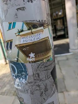 The sticker depicts a photograph of a building's balcony, from which a white banner is hung, displaying the text '#REDUSER-HUSLEIA!'. Below the balcony, a string of colorful lights is visible. At the bottom of the sticker, the word 'Rødt' is prominently displayed next to a stylized white star, which is the logo associated with the Norwegian political party, Rødt. The sticker itself is affixed to a heavily worn and layered pole in an urban setting.
