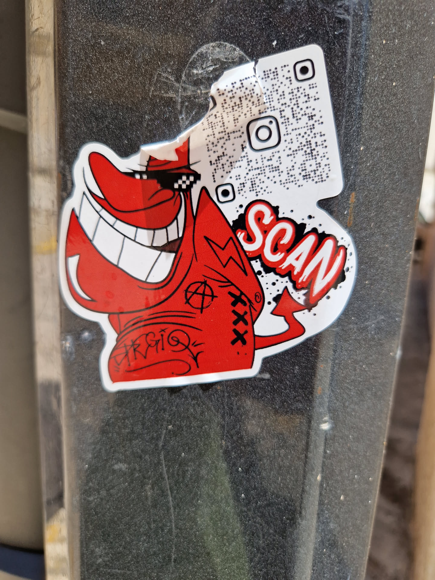 A sticker featuring a cartoon devilish character with a wide grin wearing sunglasses. The sticker also includes a QR code linking to an Instagram profile and the word 'SCAN'. The sticker is partially torn at the top.