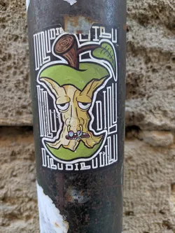 A unique sticker featuring an apple core with a whimsical, somewhat grotesque face, placed against a decorative backdrop of vertical lines. The apple core has a grumpy expression and unusual facial features, including multi-colored nostrils and a generally weathered look. The sticker is adhered to a rusty metal pole.