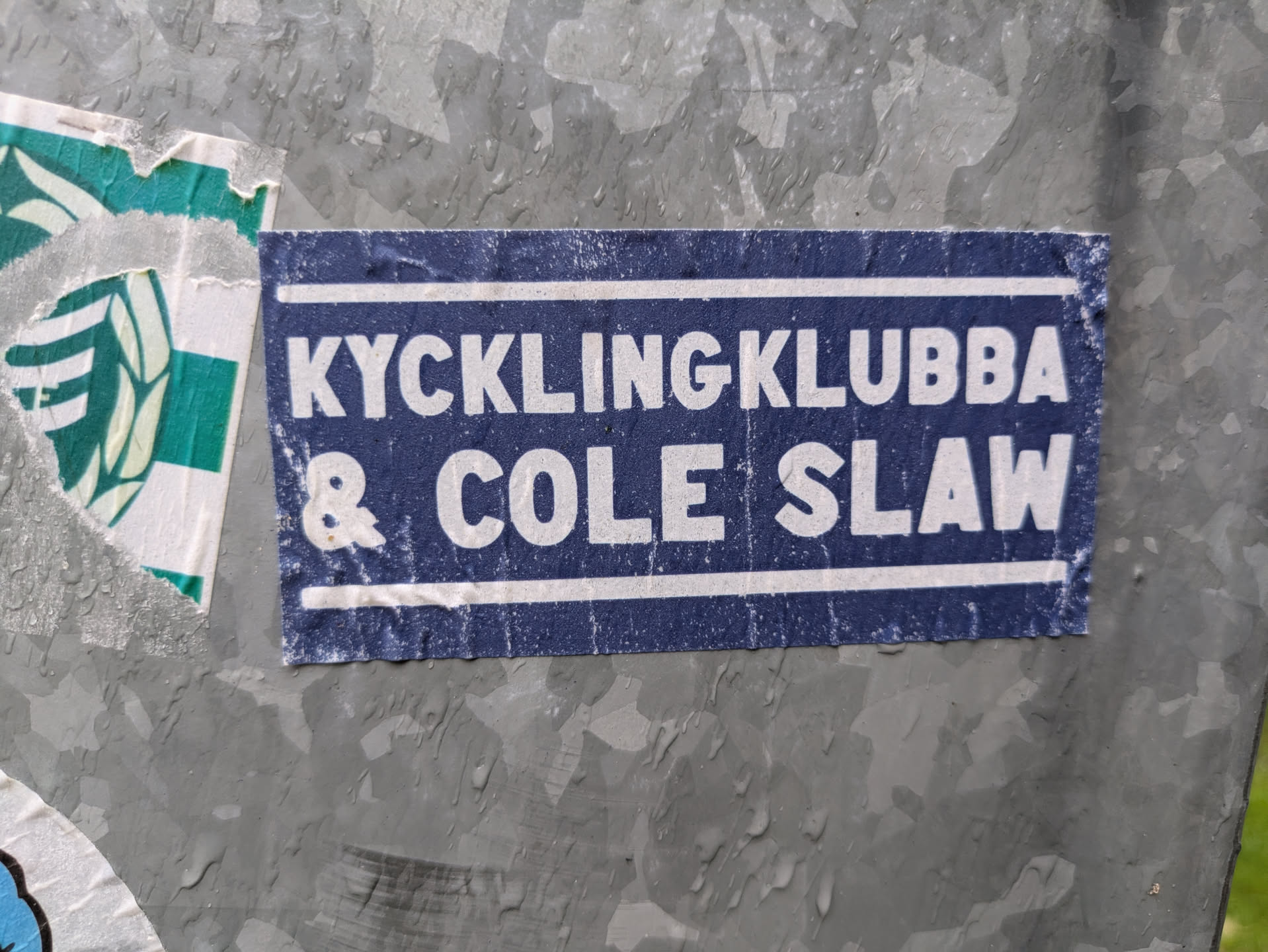 The sticker is rectangular with a dark blue background and prominent white text. It features two horizontal white lines, one near the top edge and one near the bottom edge, framing the text. The text 'KYCKLINGKLUBBA' is on the top line, and '& COLE SLAW' is on the bottom line, both in a bold, sans-serif font. The sticker shows signs of wear and tear, with some white specks and faded areas, particularly along the edges, suggesting it has been exposed to elements. It is affixed to a textured, possibly galvanized, metal surface that appears wet or has water droplets.