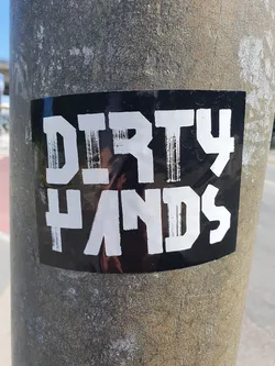 A black and white sticker with the words "Dirty Hands" in a bold, stylized font. The sticker is affixed to a grey pole.