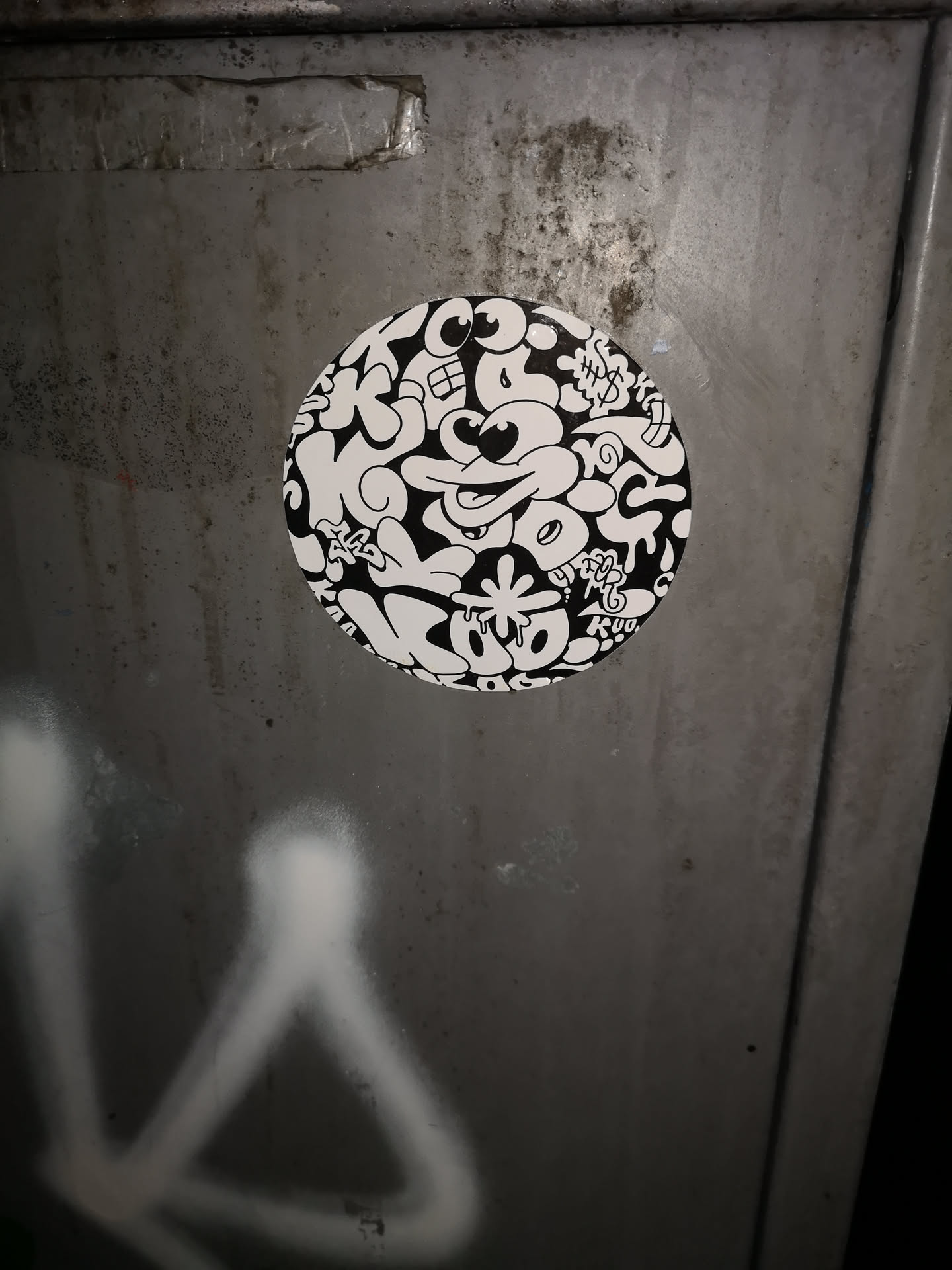 A circular, black and white sticker featuring a dense, intricate pattern of interconnected cartoonish elements. The design is a chaotic mash-up of various eyes (some wide open, some squinting), mouths (some smiling with teeth, some open), and abstract, blob-like shapes that intertwine. Elements resembling dripping paint or slime are integrated throughout. Dollar signs ($) are visible in the upper right quadrant, and a star-like shape with drips appears towards the bottom. The overall style is energetic and whimsical, with bold lines creating a strong contrast between the black and white areas.