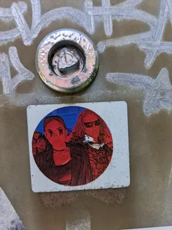 A rectangular sticker with rounded corners, featuring a circular graphic that depicts two figures in a highly stylized, red monochrome palette against a deep blue background. The figure on the left has straight hair and wears round glasses, while the figure on the right has long, curly hair and dark sunglasses. A third figure is partially visible in the far left background. The overall aesthetic suggests a vibrant, possibly pop-art or music-related theme, with strong contrasting colors. The sticker is affixed to a textured, possibly metallic surface with white graffiti markings.
