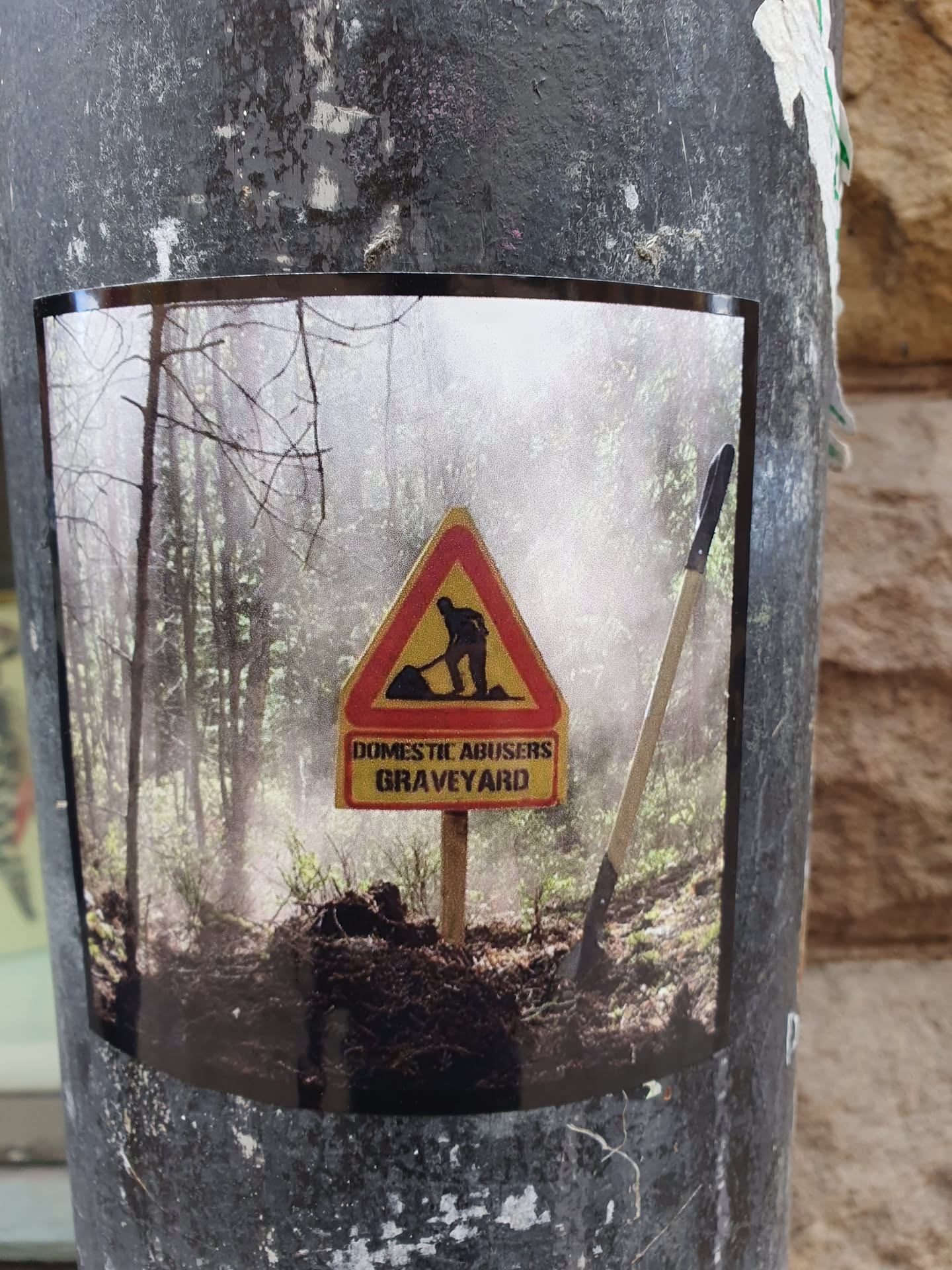 A sticker depicting a sign that reads 'Domestic Abusers Graveyard' in a forest setting.  The sign is similar to a construction or road work sign, with a figure shown digging a grave.