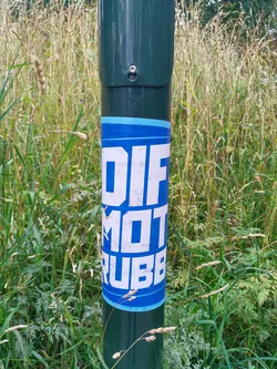 A blue and white sticker with the words "DIFF MOT RUBBER" on a dark green pole in a grassy area. The sticker shows some signs of wear.