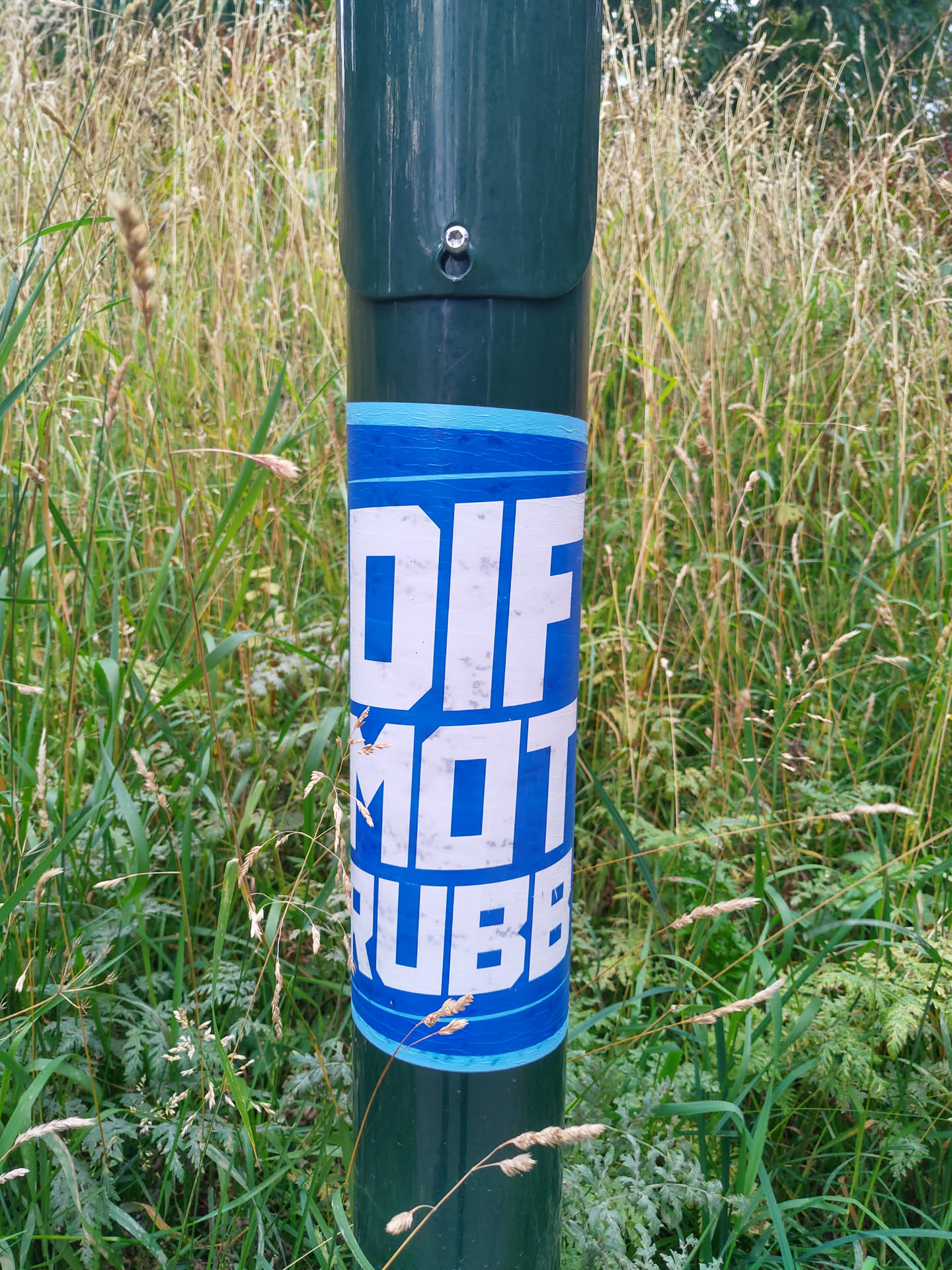 A blue and white sticker with the words "DIFF MOT RUBBER" on a dark green pole in a grassy area. The sticker shows some signs of wear.