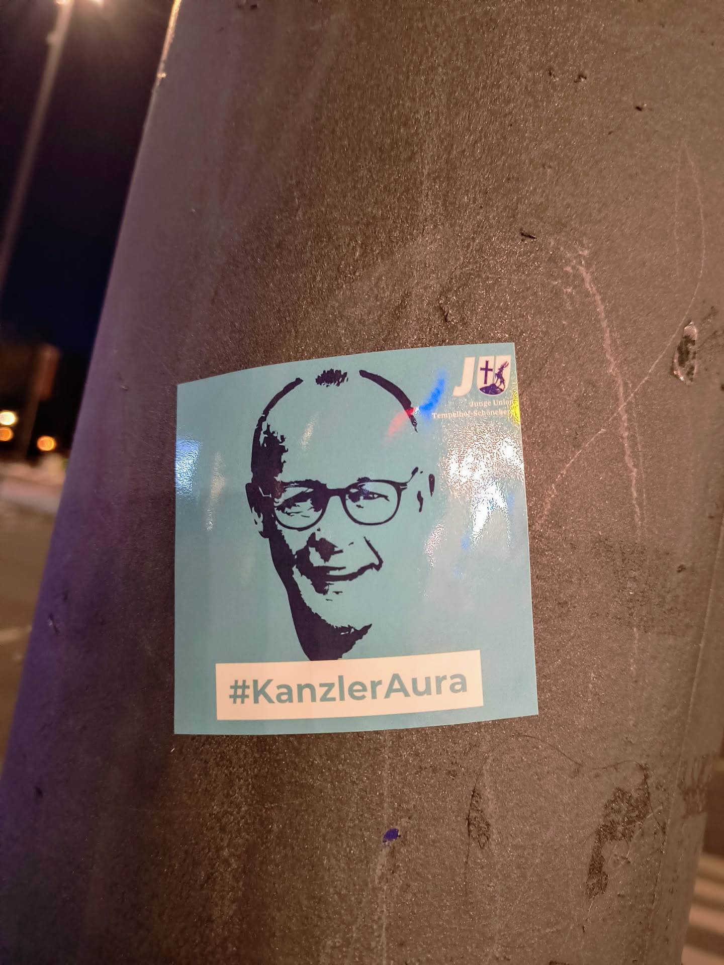 The sticker on a pole is light blue and depicts a dark blue portrait of a man wearing glasses. The sticker also includes the text "#KanzlerAura" and a logo in the top right corner, with the letters Jt.