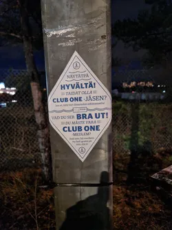 A rhombus-shaped sticker promoting Club One membership. The sticker has text in Finnish and Swedish. It is attached to a metal post outdoors at night.