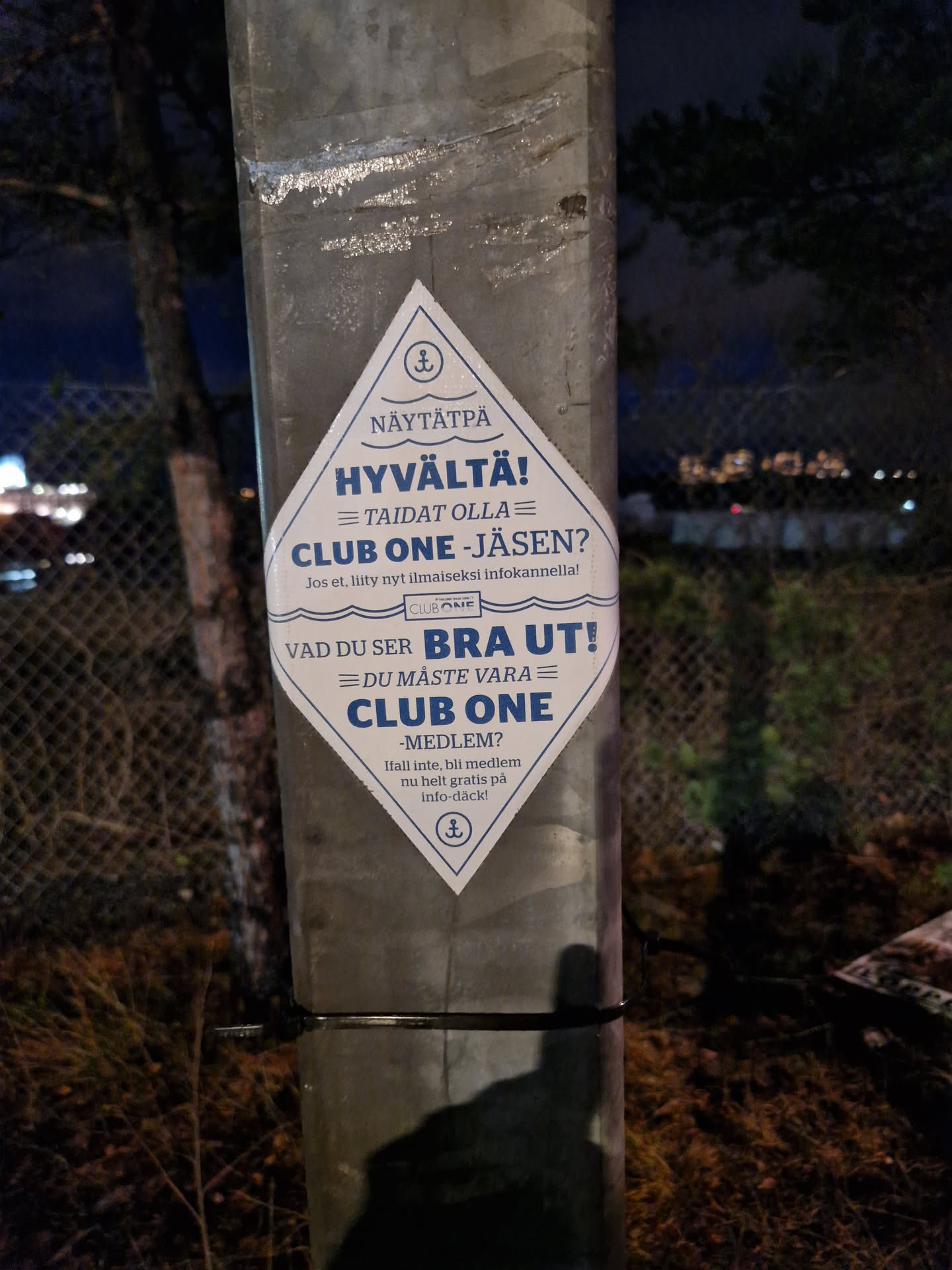 A rhombus-shaped sticker promoting Club One membership. The sticker has text in Finnish and Swedish. It is attached to a metal post outdoors at night.