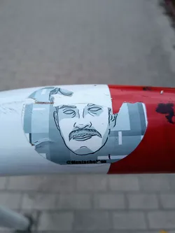 This sticker shows a stylized illustration of a man's face, prominently featuring his mustache.  The style is minimalist, using a limited color palette of blues and grays.  The sticker itself is partially damaged, with some tearing and peeling visible around its edges. The artist's Instagram handle, @MustacheFan, is printed on the lower part of the design. The sticker is affixed to a red and white pole.