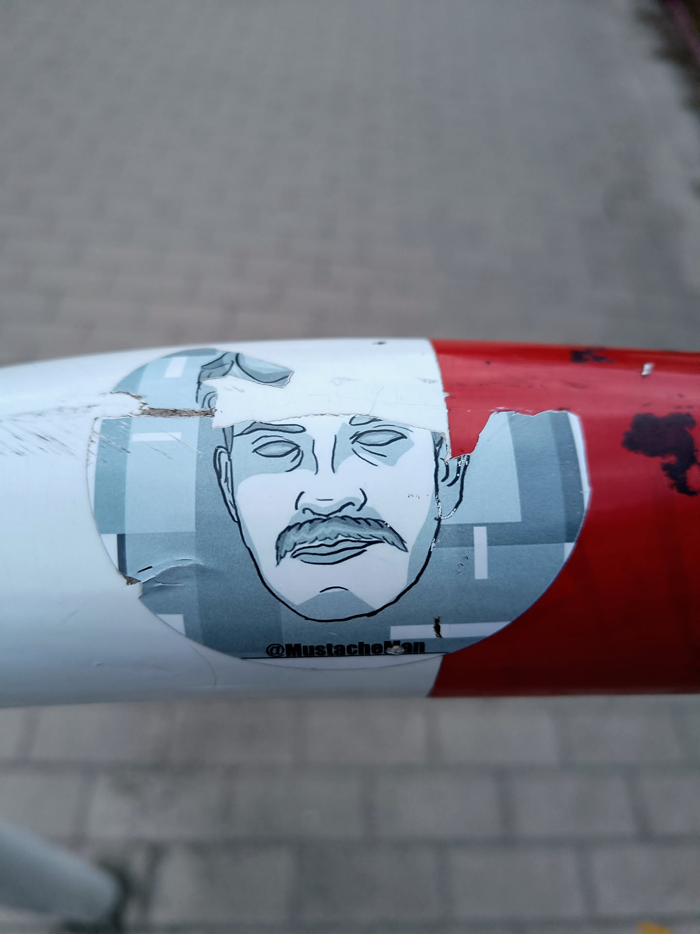 This sticker shows a stylized illustration of a man's face, prominently featuring his mustache.  The style is minimalist, using a limited color palette of blues and grays.  The sticker itself is partially damaged, with some tearing and peeling visible around its edges. The artist's Instagram handle, @MustacheFan, is printed on the lower part of the design. The sticker is affixed to a red and white pole.