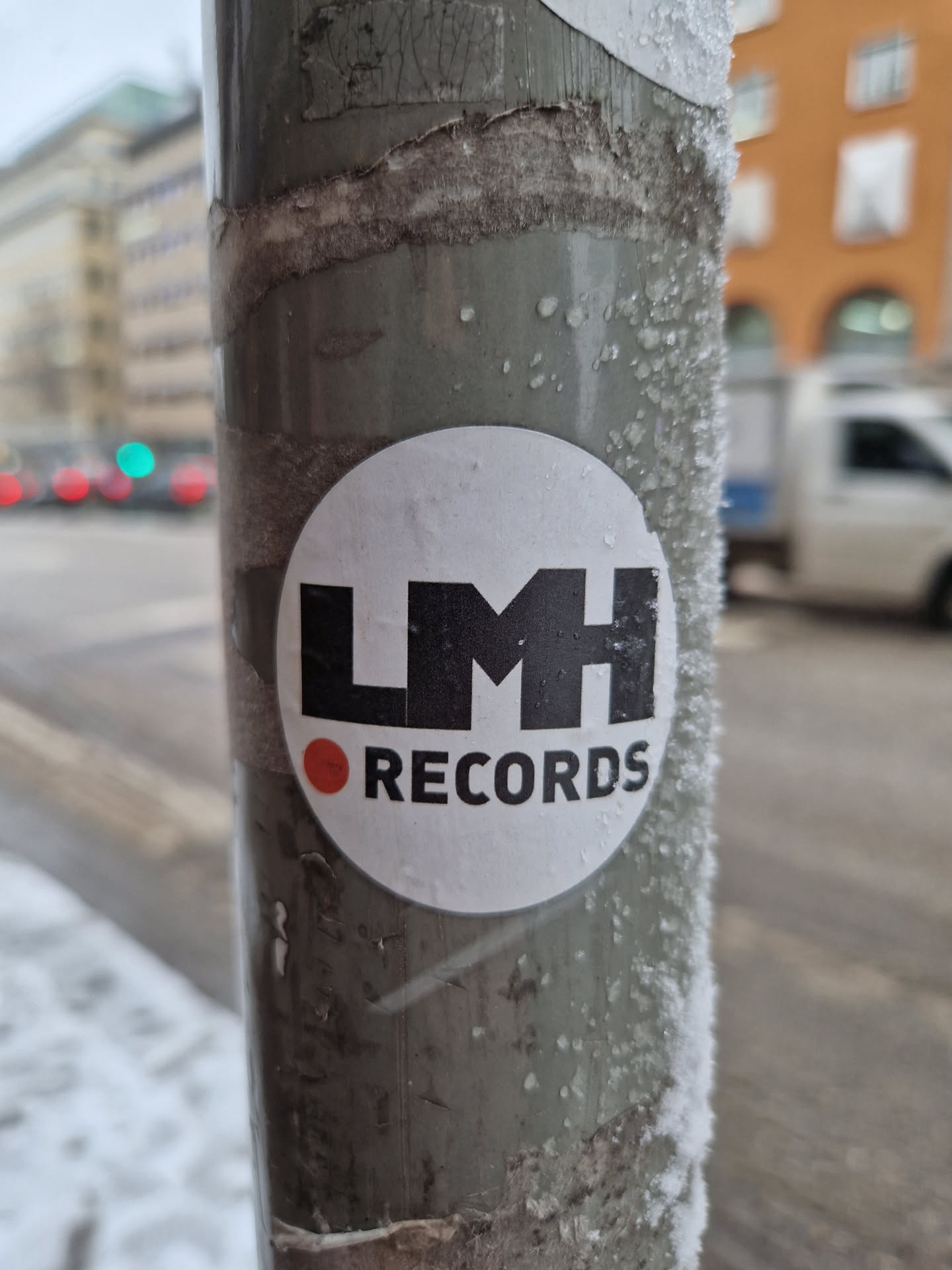 A round white sticker with the text 'LMH RECORDS' in black.  The 'LMH' is in a larger, bolder font. There is a small red dot to the left of 'RECORDS'. The sticker is affixed to a metal pole covered in a light layer of frost.