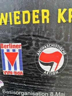 A sticker featuring a red and black flag symbol, often associated with anti-fascist movements.  The text around the symbol says "Antifaschistische Aktion".  There is also a smaller sticker visible featuring the logo for Berliner VVN-BdA.