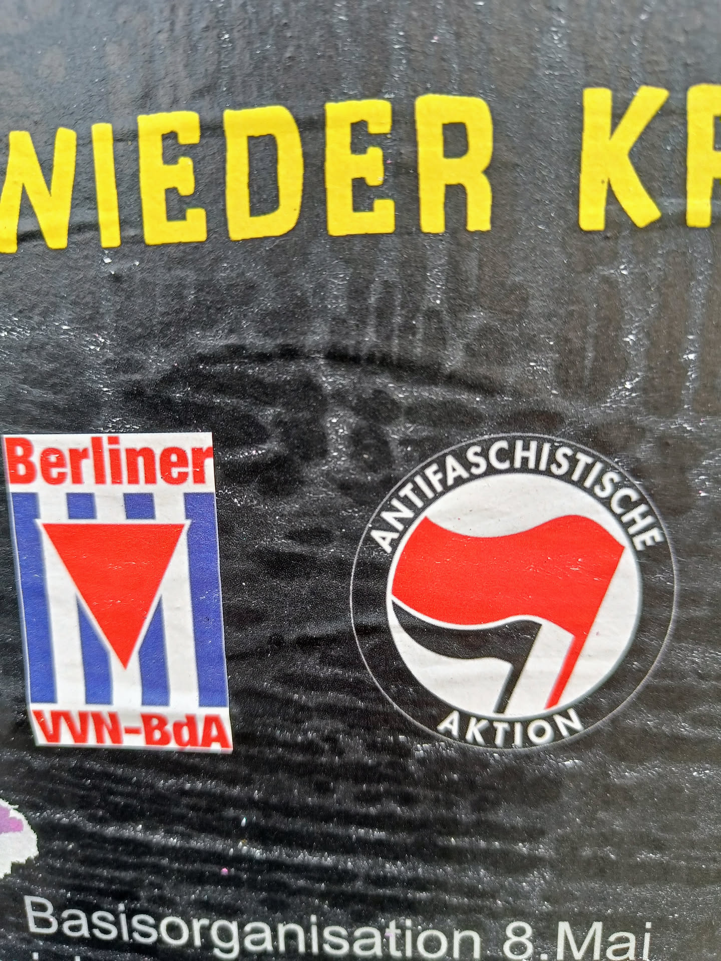 A sticker featuring a red and black flag symbol, often associated with anti-fascist movements.  The text around the symbol says "Antifaschistische Aktion".  There is also a smaller sticker visible featuring the logo for Berliner VVN-BdA.