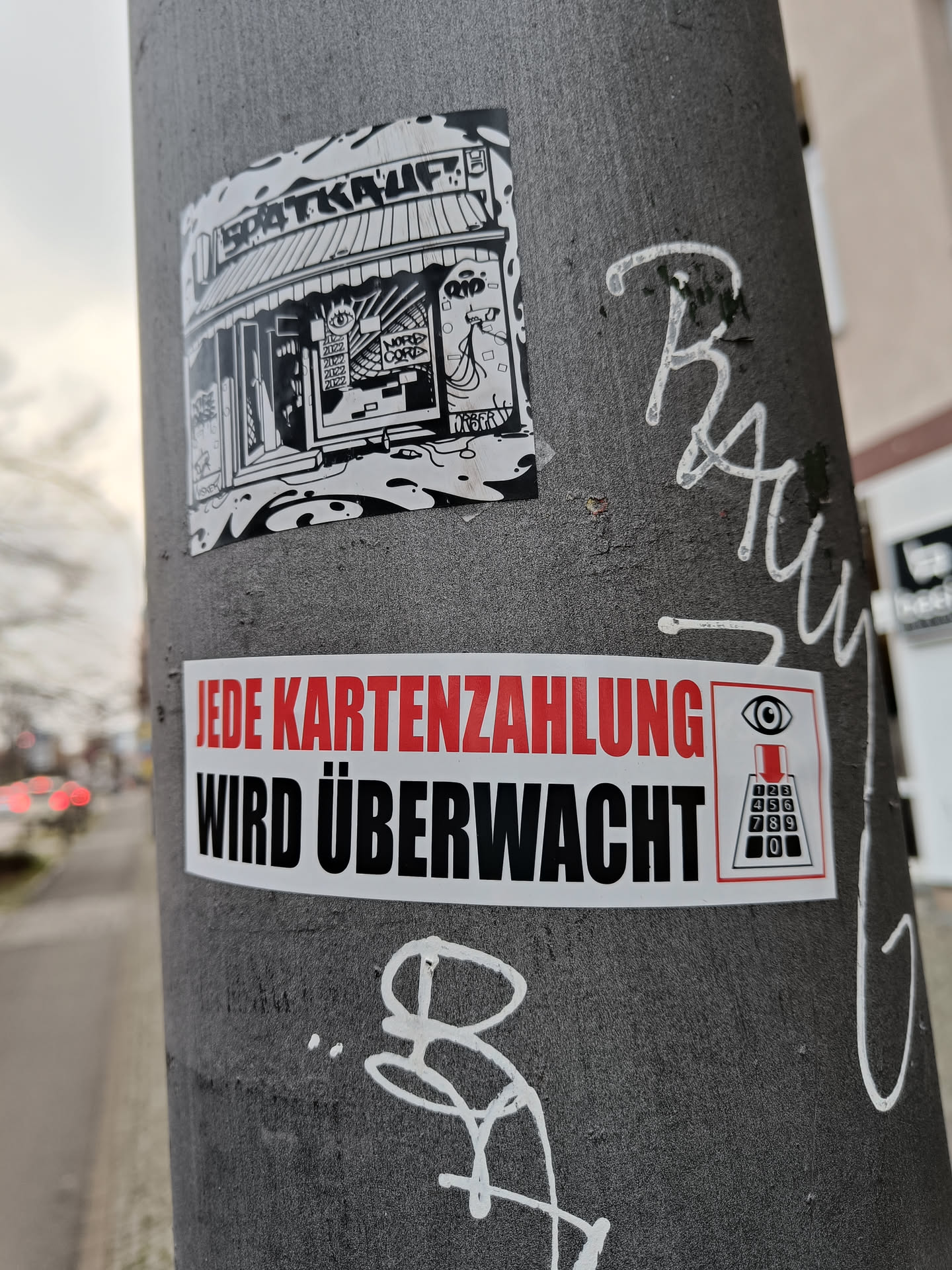 A sticker on a pole with text that says "jede Kartenzahlung wird überwacht" which translates to "every card payment is monitored". The sticker also features an illustration of a surveillance camera and a keypad. There is another sticker above it featuring an illustration of a shop called "Spätkauf".