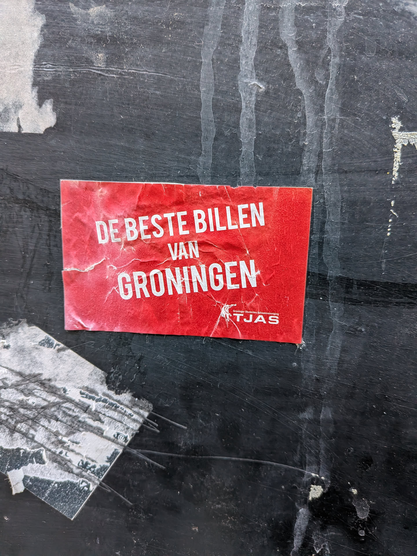 A rectangular, vibrant red sticker adhered to a dark, weathered surface. The sticker is creased and slightly torn, showing signs of age and exposure. Prominently displayed in large, white, sans-serif capital letters is the phrase 'DE BESTE BILLEN VAN GRONINGEN' (The Best Butts of Groningen). Below this, in smaller white text, it reads 'Groninger Studenten Schaatsvereniging TJAS' (Groningen Student Skating Association TJAS), accompanied by a small white silhouette logo of a person ice skating. The overall appearance is that of a well-used, outdoor promotional sticker.