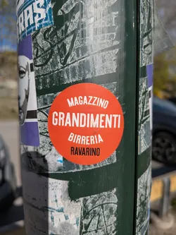 A vibrant, circular orange-red sticker prominently displayed on a dark green pole. The sticker features white, sans-serif, uppercase text arranged in four centered lines. The text reads 'MAGAZZINO', 'GRANDIMENTI', 'BIRRERIA', and 'RAVARINO'. The sticker appears relatively new and clean against the heavily weathered and stickered surface of the pole.