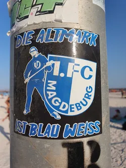 A sticker featuring a stylized illustration of a person holding a cleaning tool, possibly a brush or broom, in front of a logo that appears to be for 1. FC Magdeburg. The text 'DIE ALTMARK IST BLAU WEISS' is prominently displayed, which translates from German to "The Altmark is blue and white."