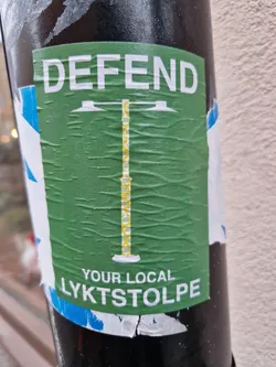Green sticker with a design of a lamppost and the text "DEFEND YOUR LOCAL LYKTSTOLPE". The sticker is attached to a pole and is slightly wrinkled.