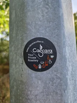 A round black sticker with Cascara Coffee Roastery branding. It features their website address, logo, and tagline, along with icons representing coffee brewing methods. The sticker is affixed to a grey metal pole.