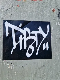 A square black sticker with white graffiti-style lettering. The text is stylized and slightly abstract. The sticker is affixed to a weathered gray surface.