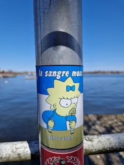 A sticker featuring Lisa Simpson from the Simpsons, holding two bottles and the text "la sangre mosa" and "since 2005". The sticker is on a metal pole near a body of water.