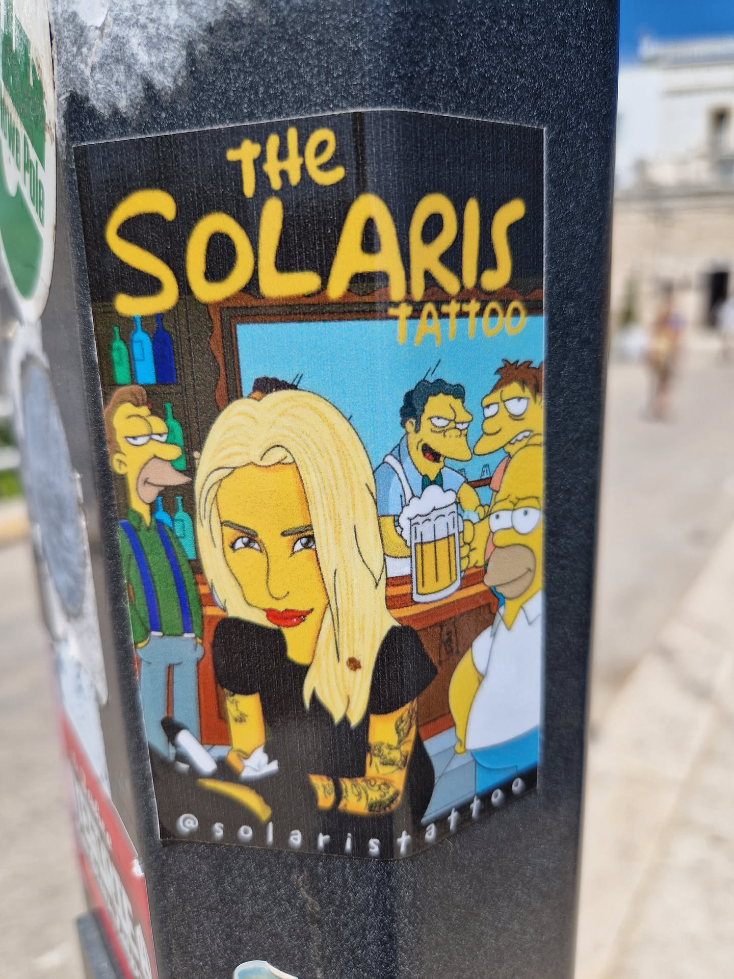 Sticker featuring a Simpsons-inspired design with a blonde woman with tattoos, surrounded by Simpsons characters in a bar scene. The sticker includes the text "The Solaris Tattoo" and the Instagram handle @solaristattoo.