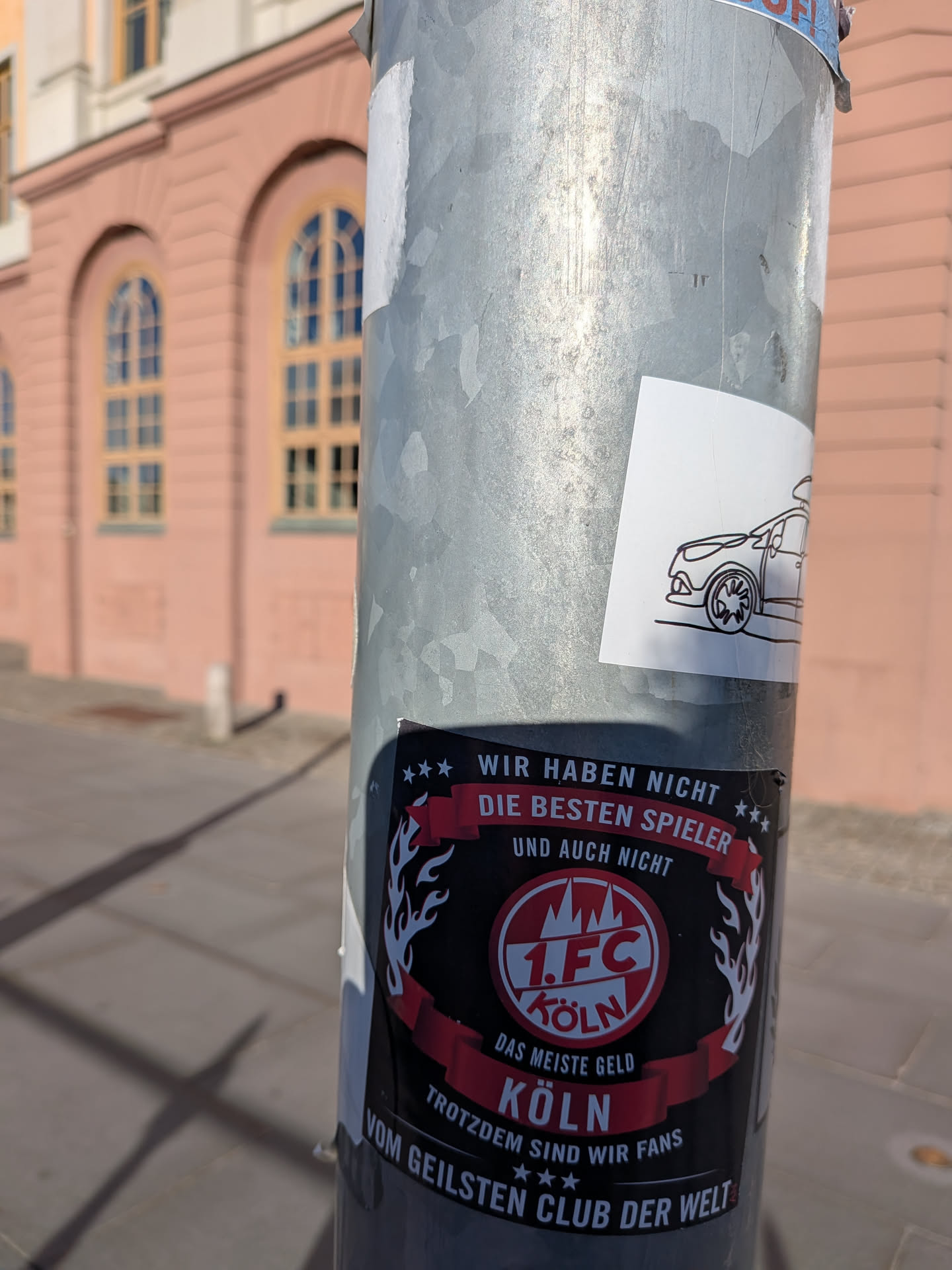 The sticker is predominantly black with red and white accents. At the center is a circular emblem featuring '1. FC KÖLN' in white text within a red circle, surrounded by a white outline and a stylized mountain graphic. Above the emblem, a red banner reads 'WIR HABEN NICHT DIE BESTEN SPIELER UND AUCH NICHT'. Below the emblem, another red banner states 'DAS MEISTE GELD KÖLN'. The bottom of the sticker features text that reads 'TROTZDEM SIND WIR FANS VOM GEILSTEN CLUB DER WELT'. The emblem is flanked by stylized white flames or wings. There is also a smaller, separate sticker above it featuring a minimalist black outline drawing of a car.