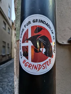 A round sticker with a red background and a black silhouette of a whale and a cross. The text on the sticker says: "The only cure for Grindadrap is you" and "#GrindStop".