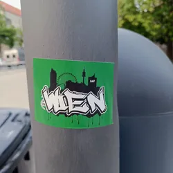 A green sticker with a graffiti-style depiction of "Wien" (Vienna in German) and a skyline featuring the Giant Ferris Wheel and the Danube Tower. The sticker has a dripping paint effect at the bottom.  There is an almost indiscernible logo on the bottom left.