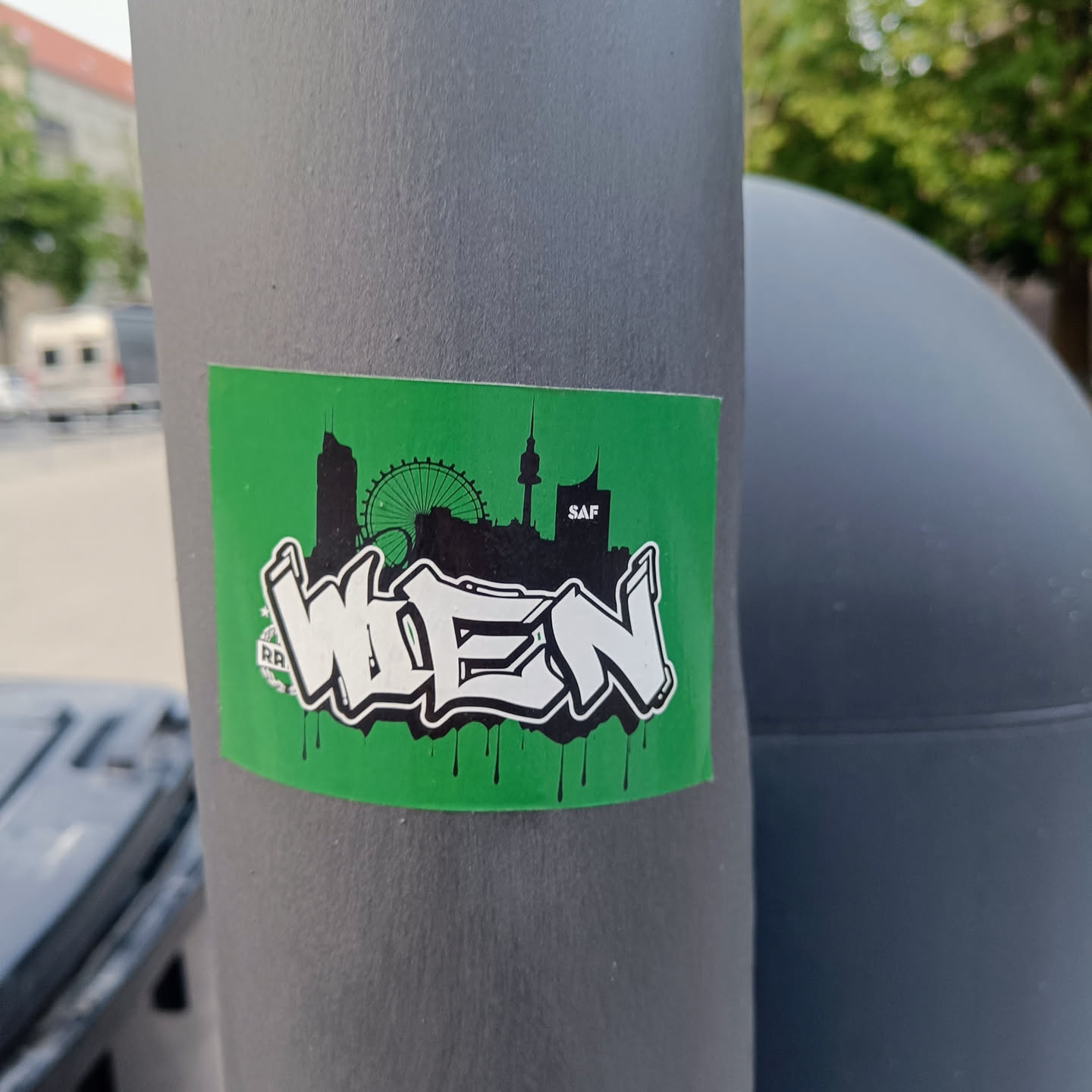 A green sticker with a graffiti-style depiction of "Wien" (Vienna in German) and a skyline featuring the Giant Ferris Wheel and the Danube Tower. The sticker has a dripping paint effect at the bottom.  There is an almost indiscernible logo on the bottom left.
