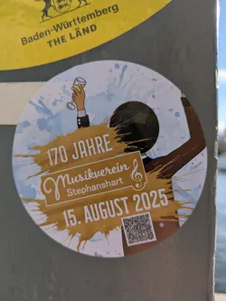 This round sticker with a white background and blue watercolor splashes, has a person celebrating something with a wine glass in his hand. The text on the sticker says that the "Musikverein Stephanshart" celebrates 170 years anniversary on August 15, 2025. A QR code in the lower right corner provides more information.