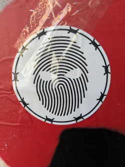 A white circular sticker is adhered to a red surface. The sticker features a design that resembles a fingerprint pattern in the center, made up of black lines. The design is encircled by a black border decorated with barbed wire illustrations. The sticker has some wrinkles.